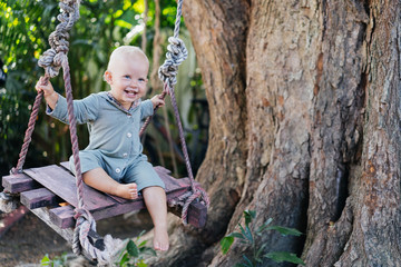 Cute happy child on a wooden swing near a tree in the summer in the tropics. Emotions and carefree childhood.