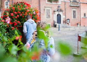  A girl in a denim jacket takes pictures, and a guy sniffs flowers in the background.