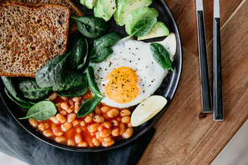 Healthy breakfast lunch at home or cafe with fried egg, avocado, toasts, beans and fresh spinach. Top down view.