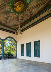 Covered terrace with decorated painted ceiling, green window shutters and marble floor fenced with metal forged fence in sunny summer day