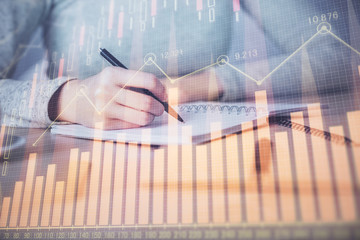 Hands writing information about stock market in notepad. Forex chart holograms in front. Concept of...