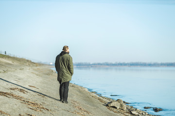 Rear view of a bearded man in a warm jacket standing against a misty river in sunny morning sunrise