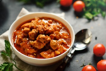 Tikka Masala. Chicken in tomato sauce in a white bowl. Spices and cherry tomatoes with parsley. Tasty chicken meat on a dark background. 