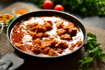 Indian food. Chicken Tikka Masala in a pan on a dark background. Appetizing meat with curry and tomato sauce, gravy with various spices. Delicious food, vertical view from the side.