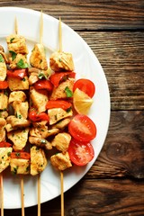 Traditional greek kebab of souvlaki with tomatoes, parsley, onions, mushrooms and paprika. top view on a white plate. Chicken meat on wooden skewers. Wooden background texture. Horizontal view 