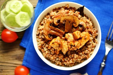 Buckwheat porridge in a white bowl. Buckwheat porridge with mushrooms and chicken breast. Wooden background and blue textile, linen napkin in blue. Top view 
