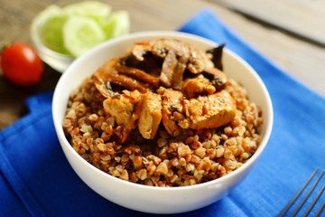 Buckwheat porridge in a white bowl. Buckwheat porridge with mushrooms and chicken breast. Wooden background and blue textile, linen napkin in blue.