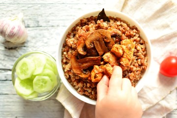 Buckwheat porridge in a white bowl on a wooden background. The child has his hands porridge with chicken breast and mushrooms. Top view, top view food. Hands in frame with food. Baby eating