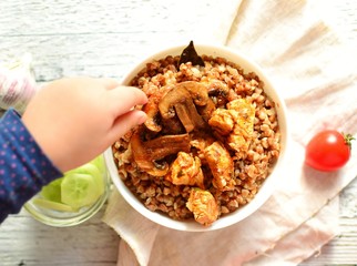 Buckwheat porridge in a white bowl on a wooden background. The child has his hands porridge with chicken breast and mushrooms. Top view, top view food. Hands in frame with food. Baby eating