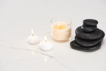 Fototapeta na wymiar burning white candles in glass near spa stones and rolled towel on marble white surface