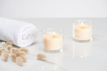 fluffy bunny tail grass near burning white candles in glass and rolled towel on marble white surface
