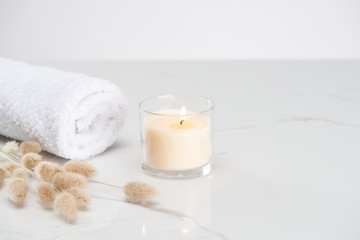 Fototapeta na wymiar fluffy bunny tail grass near burning white candle in glass and rolled towel on marble white surface