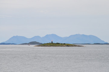 View to the small lighthouse on a small islet of Vega archipelago in Norwegian sea against the background of Gullsvågfjellet Mountain on Vega island on cloudy summer day