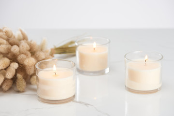 selective focus of fluffy bunny tail grass and burning white candles in glass glowing on marble...