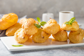 Profiterole or gougeres or eclair with custard cream filling served for breakfast on a white plate with tea on a concrete table, view from above, close-up
