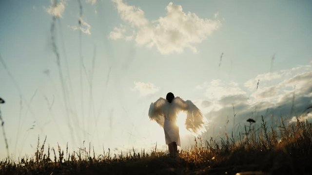 White angel and sun. Brunette girl in a white dress raises angel wings up, looks at the sun