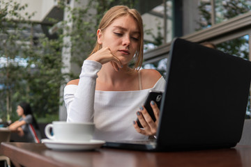 Modern business woman is reading incoming sms message on smartphone or watching broadcasting online on modern mobile phone while sitting in caffe with a laptop. Female student sitting in coffee.