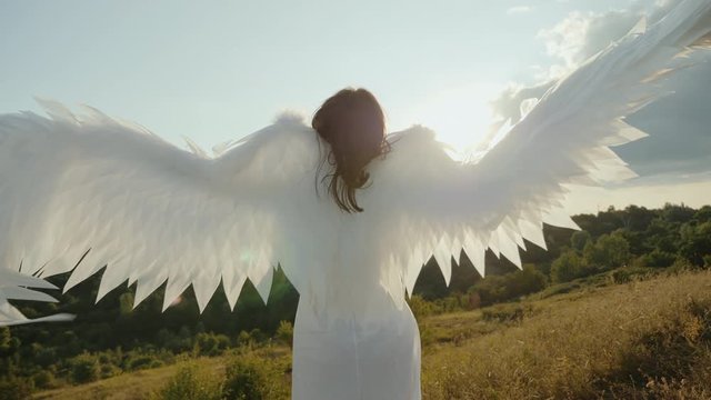 Angel with outstretched wings looks at the sun. Girl in a white dress raises angel wings up, slow-motion shot