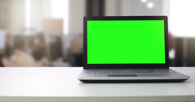 laptop computer with blank green screen on the table in office with people working in blurred background