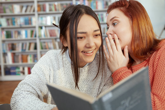 Young female students whispering quietly while studying at the library together. Asian young woman laughing while gossiping with her best friend at the college - Powered by Adobe