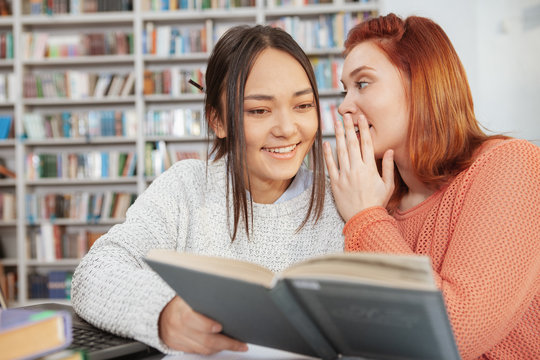 Young women studying together at the library, whispering while reading a book together. University, diversity concept - Powered by Adobe