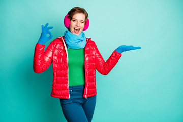 Photo of funny lady hold open arm novelty product cool offer show okay symbol wink eye wear casual red overcoat blue scarf pink ear covers jumper pants isolated teal color background