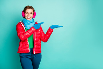 Photo of pretty lady sales manager hold open arm novelty product indicate finger wear casual red overcoat blue scarf pink ear covers jumper pants isolated teal color background
