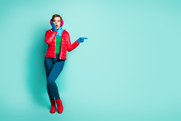 Full body photo of pretty lady indicate fingers empty space showing sale prices hand on cheek wear red overcoat blue scarf pink ear muffs pants shoes isolated teal color background