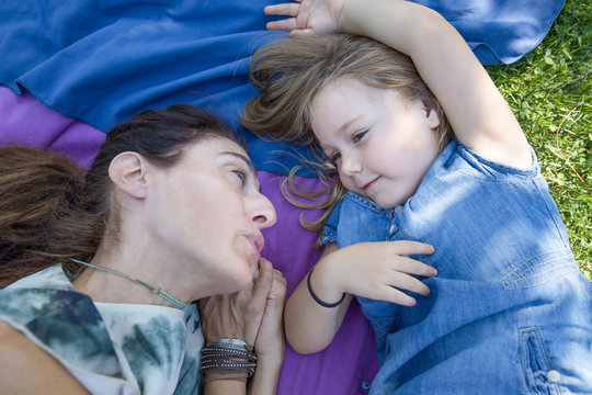 woman and four years old blonde girl lying on towels in the green grass of park with thoughtful or imagining face
