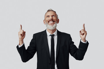 Happy mature man in full suit pointing copy space and smiling while standing against grey background