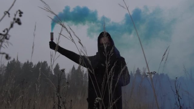 Rebellious guy wearing black clothes and face mask holding blue smoke grenade standing in the field slow motion shot.