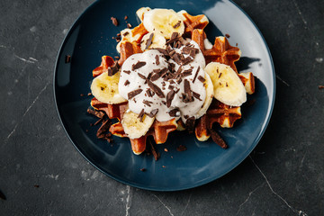 Viennese waffles on a plate, with a crumb of chocolate and slices of banana on a dark textured background.