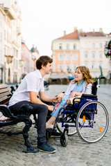 Pretty young smiling woman in the wheelchair and handsome man on the bench looking each other in love, walking in the old city center