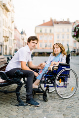 Young man and woman in wheelchair have fun and smiling on the bench outside. Old city street, love and disability concept