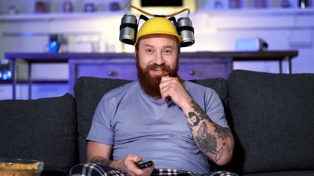 Funny bearde man with beer helmet sitting on the sofa at home and watching tv. Switching channels.