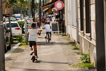 A little boy drives an e-scooter, in the background a boy with a bike, sunny day, summer weather, warm colors