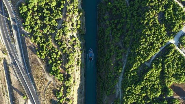 The boat goes along the Corinth Canal, surrounded by green trees. Aerial view. 