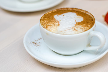 hold ceramic white with cappuccino above wooden plate and rustic table. Milk foam on top in heart shape