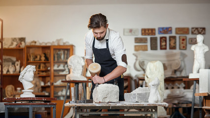 Bearded craftsman works in white stone carving with a chisel. Creative workshop with works of art.