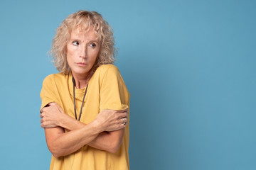 Senior woman with blonde hair in yellow shirt shaking from cold angrily looking in camera over blue background
