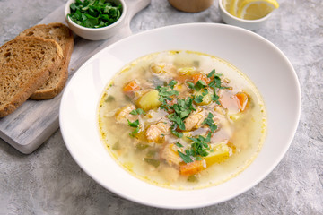 Fish soup made of salmon and vegetables in white bowl, served with bread toasts, parsley, lemon on textured grey table. Vegetarian and healthy food rich with Omega 3.