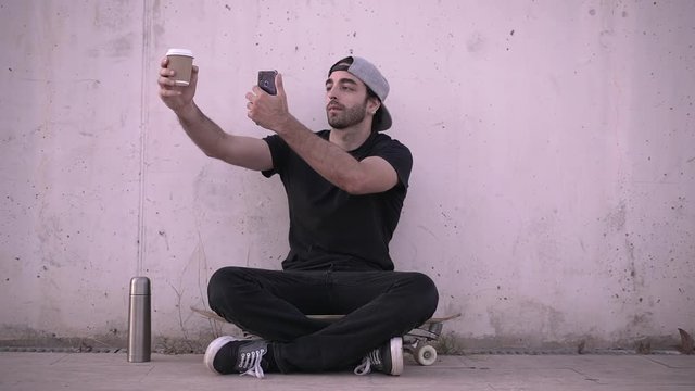 Man sitting on skateboard using phone and drinking coffee