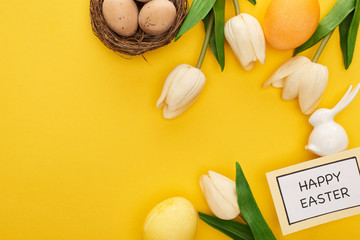 top view of tulips, greeting card with happy Easter lettering, easter bunny and eggs in nest on yellow colorful background