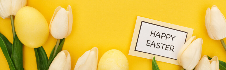 top view of tulips and painted Easter eggs near card with happy Easter lettering on colorful yellow background, panoramic shot