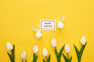 top view of tulips, greeting card with happy Easter lettering near Easter white bunnies on yellow colorful background