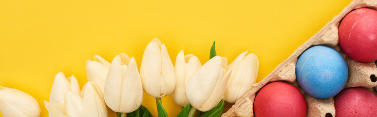 top view of tulips and multicolored painted Easter eggs in cardboard container on colorful yellow background, panoramic shot