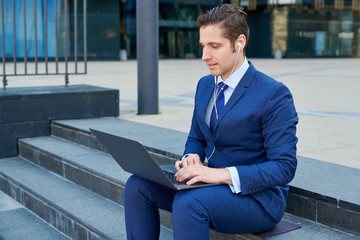 Happy sucessfull young businessman working on modern laptop, using earphones and sitting on stairs outside skyscapers