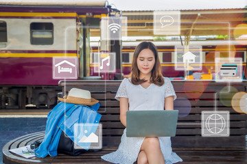 Beautiful Asian woman in  white dress siting on wooden bench with jean jacket cover her backpack using laptop with technology icon around on her at train station