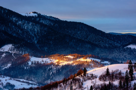 winter countryside scenery at dawn. landscape with spot of first light on snow covered hill. dark coniferous distant forest in shade of the mountain. cold weather.