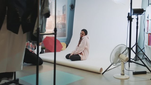 Young caucasian photographer taking photos of pretty african model sitting on the floor in studio photo shoot. There is modern lightning equipment.